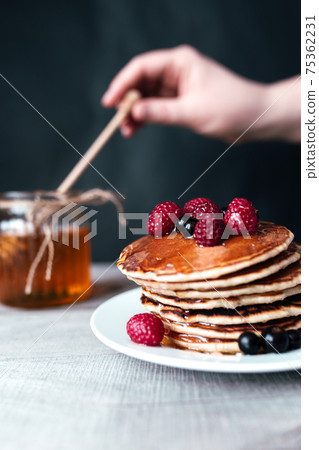 Pancakes with berries and honey on white plate, hand holding spoon in jar 75362231
