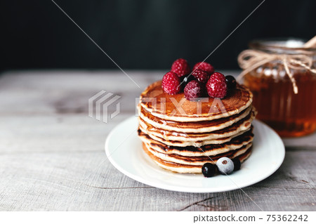 Juicy pancakes with berries and honey on a white plate, spoon, jar, wooden table 75362242