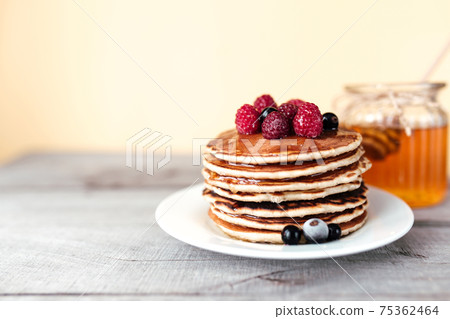 Juicy pancakes with berries and honey on a white plate, spoon, jar, wooden table 75362464