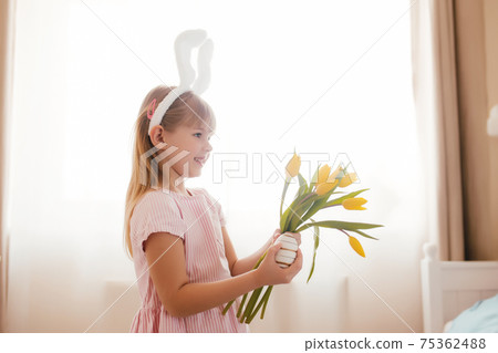 Easter concept. Girl in bunny ears and pink dress holding yellow tulips and egg Easter concept. Girl in bunny ears and pink dress holding yellow tulips and egg 75362488