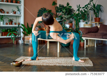 Young sporty woman practicing yoga at home doing twisting variation exercise of goddess pose Young sporty woman practicing yoga at home doing twisting variation exercise of goddess pose 75363877