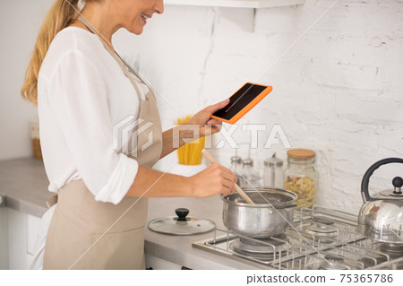 Woman in apron cooking and watching something online Woman in apron cooking and watching something online 75365786