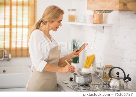 Woman in apron cooking and watching something online Woman in apron cooking and watching something online 75365787