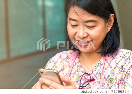 Portrait Asian middle-aged woman, close up Asian woman with smiling face while looking at her cellphone, wearing casual, hang eyeglasses on tee shirt's neck. Asian woman with short black hair. 75366234