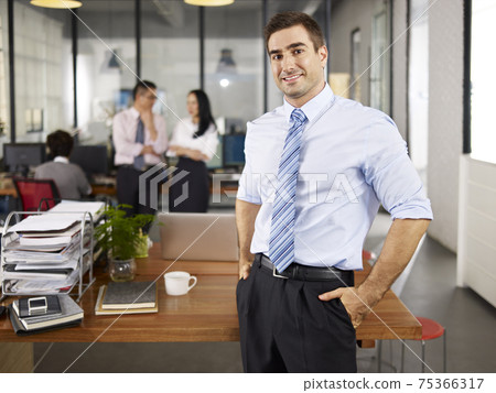 successful caucasian expat businessman standing in office local colleagues talking in background 75366317