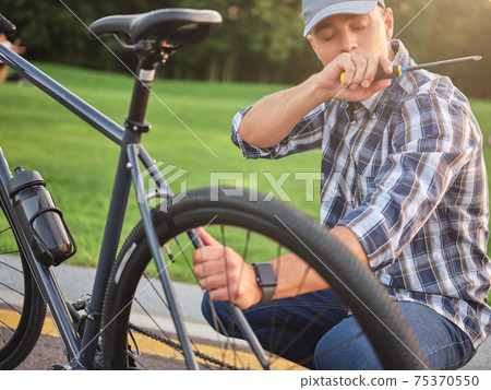Young caucasian man holding a screwdriver while checking and repairing his bike in the public park 75370550