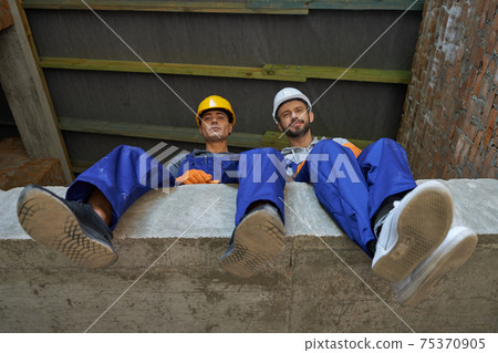 Two attractive young builders in blue overalls and hard hats looking down at camera, sitting on the concrete floor, resting while working on house construction Two attractive young builders in blue overalls and hard hats looking down at camera, sitting on the concrete floor, resting while working on house construction 75370905