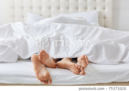 Horizontal shot of feet of couple under white bedclothes in bed at bedroom, enjoying good rest at home and calm atmosphere. Female and male relaxing together at weekend. Pomantic couple in love. 75371028