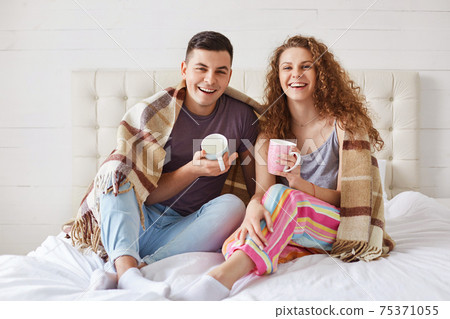 Positive young family couple wear pyjamas, drink coffee in morning at bedroom, enjoy starting new day. Affectionate female and make enjoy togetherness at cozy domestic atmosphere. Family concept Positive young family couple wear pyjamas, drink coffee in morning at bedroom, enjoy starting new day. Affectionate female and make enjoy togetherness at cozy domestic atmosphere. Family concept 75371055