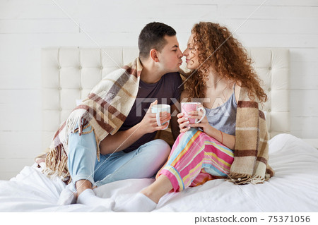 Indoor shot of happy female and male have romantic relationships, drink coffee or tea, covered with plaid in bedroom. Romantic couple enjoying spending leisure together. Relationship concept. 75371056