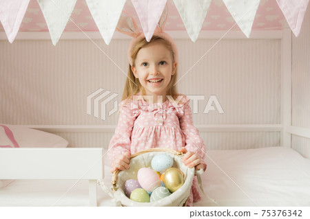 Little girl wearing bunny ears and holding a basket of Easter eggs. Happy Easter. Preparing family for Easter. Easter bunny ears.Smiling face Little girl wearing bunny ears and holding a basket of Easter eggs. Happy Easter. Preparing family for Easter. Easter bunny ears.Smiling face 75376342