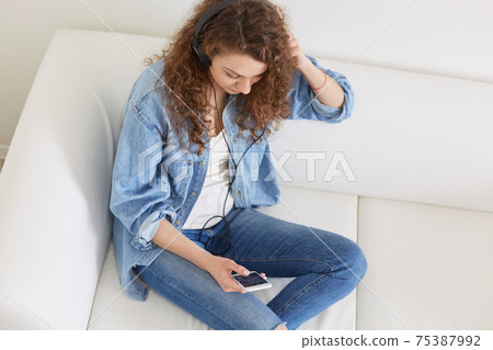 Indoor shot of attractive lady with headphones, listening to music, top view of concentrated relaxed model, sitting on sofa, enjoying her free time, wearing casual clothes. People and rest concept. 75387992