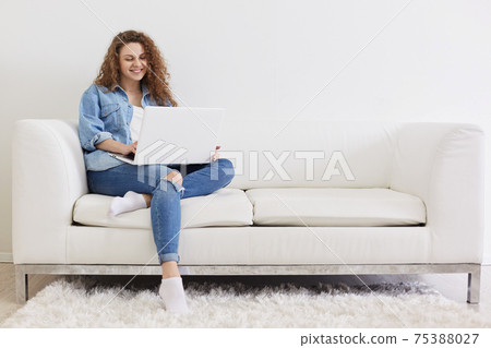 Photo of charming nice satisfied lady with curly hair sitting on white comfortable sofa, using laptop for online working, using wireless Internet, being in light interior room, looks happy and glad. 75388027