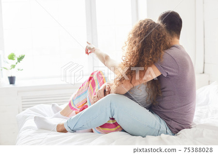 Lovely girlfriend and boyfriend dressed casually, enjoy intimacy and togetherness, pose on bed in bedroom, look at window, have pleasant talk, express good feeling to each other. Bed time concept Lovely girlfriend and boyfriend dressed casually, enjoy intimacy and togetherness, pose on bed in bedroom, look at window, have pleasant talk, express good feeling to each other. Bed time concept 75388028