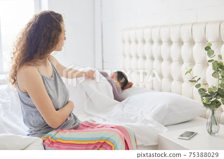 Curly beautiful woman in pyjamas sits on side of bed, dressed in casual pyjamas, tries to awake her husband, have problems with relatioships, pose in cozy bedroom, Family and sleeping concept 75388029