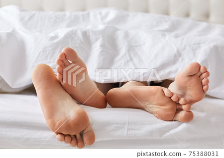Female`s and male`s bare feet have rest under white blanket, recreat together, feels relaxation. Unrecognizable family couple sleep in comfortable bed. Bed time and good rest concept 75388031