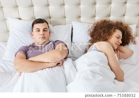 Horizontal shot of lovely faily couple have rest in bed, enjoy domestic atmosphere, like spend time in comfortable bed, Serious man keeps hands crossed, feels bored while girlfriend is sleeping Horizontal shot of lovely faily couple have rest in bed, enjoy domestic atmosphere, like spend time in comfortable bed, Serious man keeps hands crossed, feels bored while girlfriend is sleeping 75388032