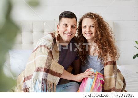 Close up portrait of romantic couple in love sitting under warm plaid in bedroom, embrace each other and have positive facial expressions, enjoy togetherness, dressing casually, looking at camera. Close up portrait of romantic couple in love sitting under warm plaid in bedroom, embrace each other and have positive facial expressions, enjoy togetherness, dressing casually, looking at camera. 75388033