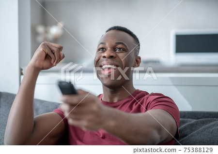 A dark-skinned young man watching tv and looking excited 75388129