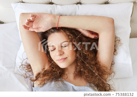 Horizontal portrait of sleepy cute female with curly hair, pleasant appearance, feels relaxed and sees good dreams, lies in comfortable bed, enjoys calm domestic atmopshere. Sleeping concept Horizontal portrait of sleepy cute female with curly hair, pleasant appearance, feels relaxed and sees good dreams, lies in comfortable bed, enjoys calm domestic atmopshere. Sleeping concept 75388132