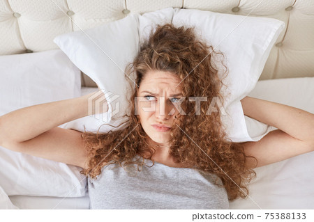 Pretty discontent female with curly covers ears with pillow, lies in bed, has insomnia, cant fall asleep because of noise, has sleepless night, looks in displeasure aside. Stop this annoying sound Pretty discontent female with curly covers ears with pillow, lies in bed, has insomnia, cant fall asleep because of noise, has sleepless night, looks in displeasure aside. Stop this annoying sound 75388133