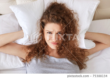 Depressed frustrated female covers ears with pillow while lies in bed, can`t stand someone`s snoring, can`t get asleep, suffers from insomnia. Curly young woman has some problems with sleep. Depressed frustrated female covers ears with pillow while lies in bed, can`t stand someone`s snoring, can`t get asleep, suffers from insomnia. Curly young woman has some problems with sleep. 75388134