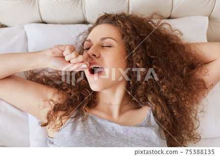 Indoor shot of pretty woman yawns in bed as wakes up early in morning, has curly bushy hair, starts new day, has sleepy expression, wants to stay in bed for long time. Yawning female in bedroom Indoor shot of pretty woman yawns in bed as wakes up early in morning, has curly bushy hair, starts new day, has sleepy expression, wants to stay in bed for long time. Yawning female in bedroom 75388135