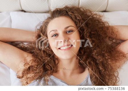 Top view of cheerful young female with curly bushy hair has pleasant smile as lies on comfortable bed, enjoys starting new day or good morning, likes relax in bed. Bed time and rest concept. Top view of cheerful young female with curly bushy hair has pleasant smile as lies on comfortable bed, enjoys starting new day or good morning, likes relax in bed. Bed time and rest concept. 75388136