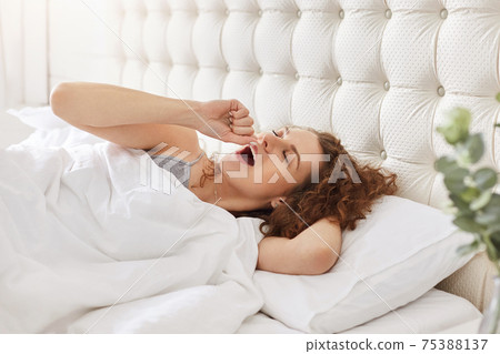 Morning and awakening concept. Young woman yawns as lies in comfortable bed, wants to sleep late at night, rests in spacious bedroom. Peaceful atmosphere. People and relaxation concept 75388137