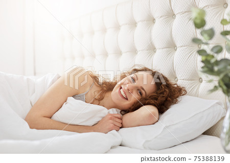 Smiling lovely young woman wakes up in morning, has gladful expression, lies on white bedclothes in bed, poses at camera with satisfied expression. People, home, morning time and rest concept Smiling lovely young woman wakes up in morning, has gladful expression, lies on white bedclothes in bed, poses at camera with satisfied expression. People, home, morning time and rest concept 75388139