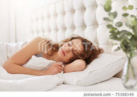 Candid shot of pleased young woman enjoys good sleep in bedroom, lies in comfortable bed, has appealing appearance, keeps eyes closed, lies under white bedclothes. Beautiful female has rest. Candid shot of pleased young woman enjoys good sleep in bedroom, lies in comfortable bed, has appealing appearance, keeps eyes closed, lies under white bedclothes. Beautiful female has rest. 75388140