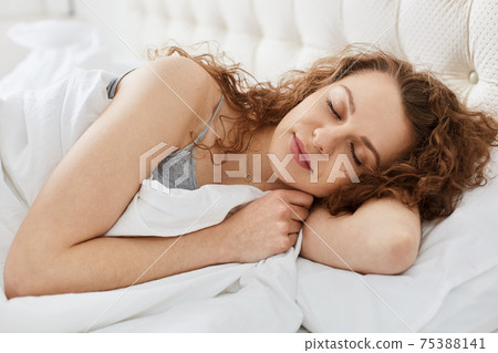 Close up morning portrait of attractive young sensual woman laying on white bed close her eyes, female has long curly hair and pretty face, sleeping with calm facial expression. Recreation concept. Close up morning portrait of attractive young sensual woman laying on white bed close her eyes, female has long curly hair and pretty face, sleeping with calm facial expression. Recreation concept. 75388141