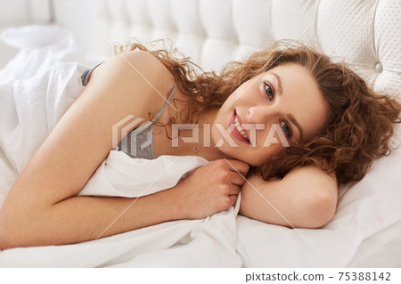 Horizontal portrait of pleased happy woman with cheerful expression, lies on comfortable bed, feels relaxed after good rest, enjoys in comfortable nest. Cute female napes in bedroom. Horizontal portrait of pleased happy woman with cheerful expression, lies on comfortable bed, feels relaxed after good rest, enjoys in comfortable nest. Cute female napes in bedroom. 75388142