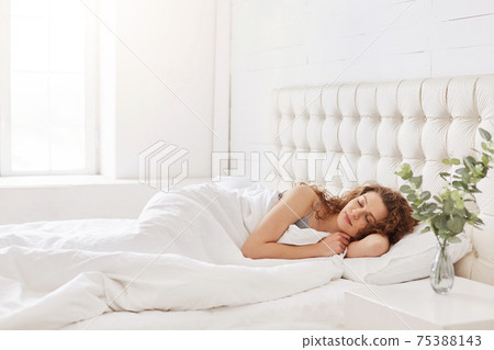 Horizontal shot of relaxed carefree female under white bedclothes in bed at bedroom, sees pleasant dreams, keeps eyes closed, enjoys good rest at home and calm atmosphere. People and rest concept Horizontal shot of relaxed carefree female under white bedclothes in bed at bedroom, sees pleasant dreams, keeps eyes closed, enjoys good rest at home and calm atmosphere. People and rest concept 75388143