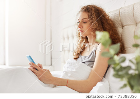 Sideways portrait of serious female with attractive look, sits under white soft bedclothes, reads news online on modern smart phone, wants to know what happened for day. People, leisure, bed time Sideways portrait of serious female with attractive look, sits under white soft bedclothes, reads news online on modern smart phone, wants to know what happened for day. People, leisure, bed time 75388356