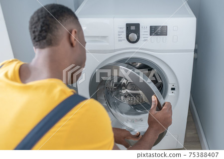 Dark-skinned service man in yellow tshirt repairing the washing machine Dark-skinned service man in yellow tshirt repairing the washing machine 75388407