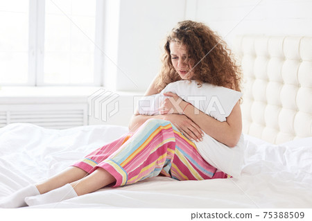 Stressed cury female wears pyjamas, holds pillow, suffers as has periods or menstruation, sits on soft bed with white bedclothes, has sorrowful facial expression. People, health problems concept Stressed cury female wears pyjamas, holds pillow, suffers as has periods or menstruation, sits on soft bed with white bedclothes, has sorrowful facial expression. People, health problems concept 75388509