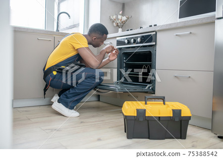 Dark-skinned service man in yellow tshirt repairing the gas stove Dark-skinned service man in yellow tshirt repairing the gas stove 75388542