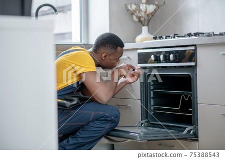 Dark-skinned service man in yellow tshirt repairing the gas stove Dark-skinned service man in yellow tshirt repairing the gas stove 75388543
