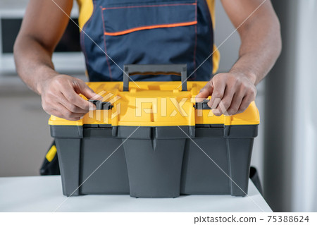 Dark-skinned service man standing in the kitchen and opening his tools box Dark-skinned service man standing in the kitchen and opening his tools box 75388624