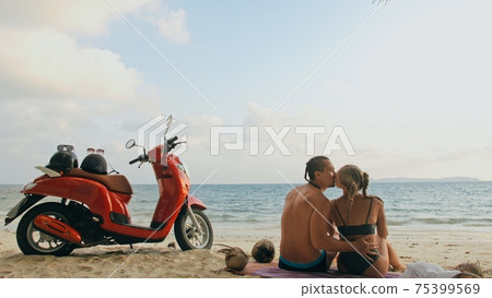 Scooter road trip. Lovely couple on red motorbike in white clothes on sand beach. People walking near the tropical palm trees, sea. Motorcycle rent. 75399569
