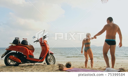Scooter road trip. Lovely couple on red motorbike in white clothes on sand beach. People walking near the tropical palm trees, sea. Motorcycle rent. Scooter road trip. Lovely couple on red motorbike in white clothes on sand beach. People walking near the tropical palm trees, sea. Motorcycle rent. 75399584