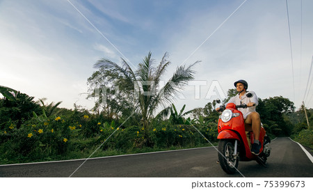 Man on red motorbike in white clothes drive on forest road trail trip. One men caucasian tourist go on scooter, nearby tropical palm tree. Man on red motorbike in white clothes drive on forest road trail trip. One men caucasian tourist go on scooter, nearby tropical palm tree. 75399673