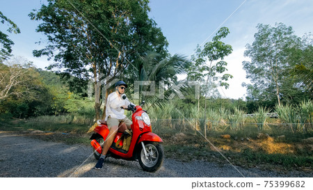 Man on red motorbike in white clothes drive on forest road trail trip. One men caucasian tourist go on scooter, nearby tropical palm tree. 75399682
