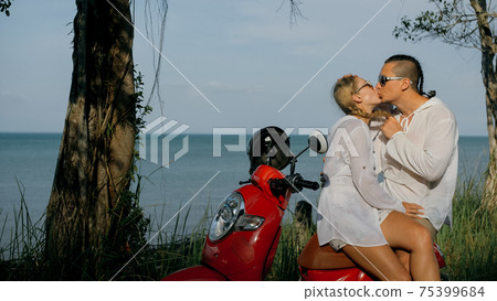 Love couple on red motorbike in white clothes on forest road trail trip. Two caucasian tourist woman and man kiss hugs sit on scooter. 75399684
