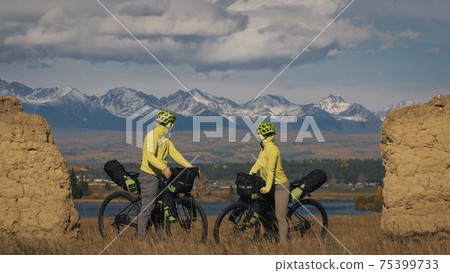 The man and woman travel on mixed terrain cycle touring with bikepacking. The two people journey with bicycle bags. Mountain snow capped, stone arch. The man and woman travel on mixed terrain cycle touring with bikepacking. The two people journey with bicycle bags. Mountain snow capped, stone arch. 75399733