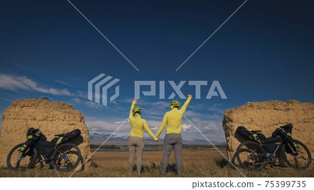 The man and woman travel on mixed terrain cycle touring with bikepacking. The two people journey with bicycle bags. Mountain snow capped, stone arch. 75399735