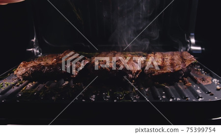 Delicious juicy meat steak cooking on grill. Prime beef fry on electric roaster, rosemary, black pepper, salt. Slow motion. Delicious juicy meat steak cooking on grill. Prime beef fry on electric roaster, rosemary, black pepper, salt. Slow motion. 75399754