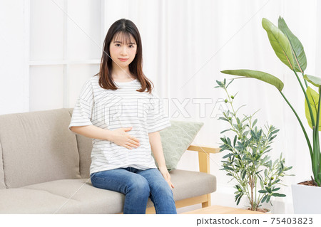 A young woman sitting on the sofa in the room and putting her hand on her belly 75403823