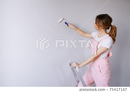 A woman in a pink jumpsuit is painting a wall. Renovation and new life for old things. A woman in a pink jumpsuit is painting a wall. Renovation and new life for old things. 75417187